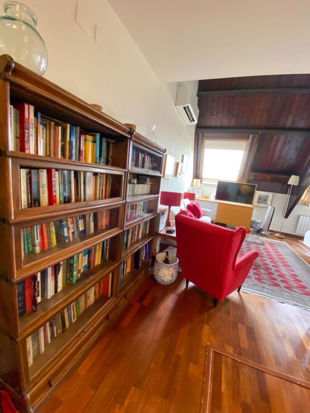 A room with wooden bookshelves filled with books, a red chair, a lamp, and a large window.