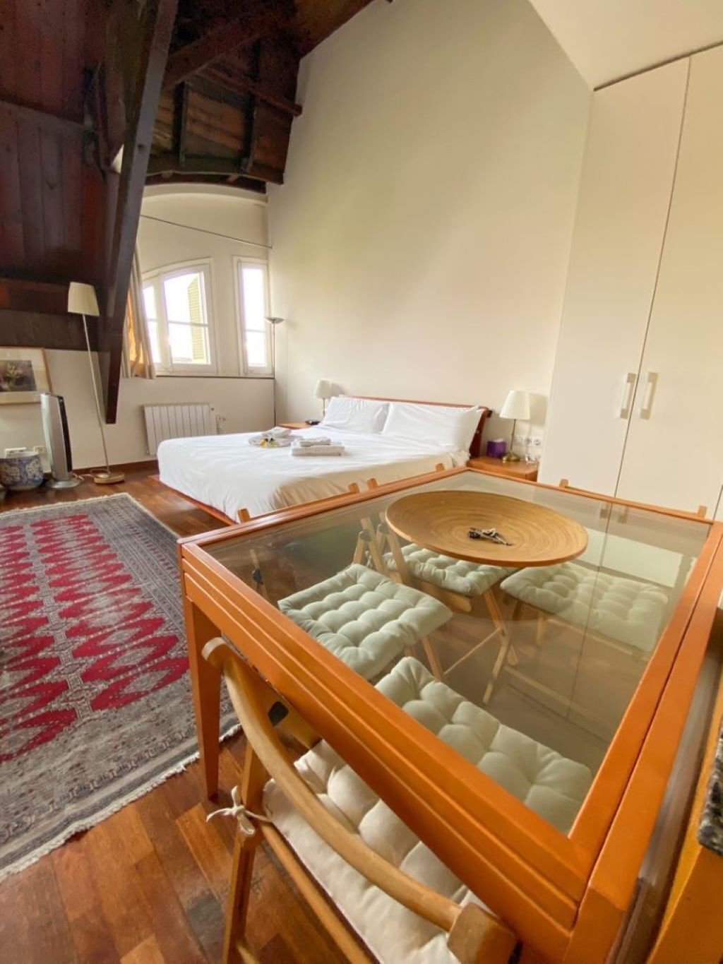 Un dormitorio con una cama grande, una mesa de vidrio con sillas acolchadas, un tapete rojo y gris, y un armario blanco.