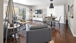 A modern living room with a grey sofa, dining table, and chairs. There is a TV mounted on the wall and a large window with blinds. - Renovated, Ground Level, 2 BR 2 BA, WiFi, Cable, Pool. 4 individuals max. (Pinehurst)