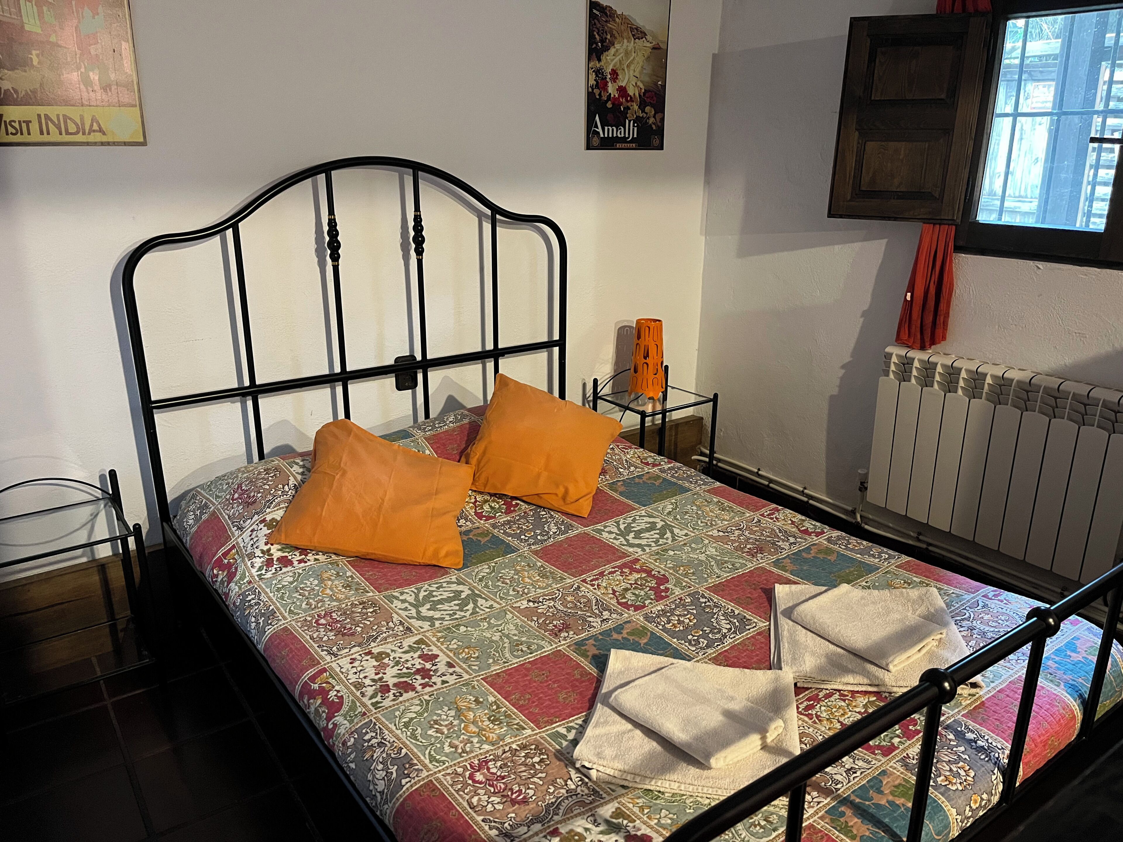 Una cama con colcha estampada, dos almohadas, una mesita de noche con lámpara y una ventana con cortinas.