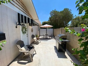 Um alpendre ao ar livre com uma churrasqueira, duas cadeiras Adirondack, uma mesa com guarda-sol e um vaso de plantas.