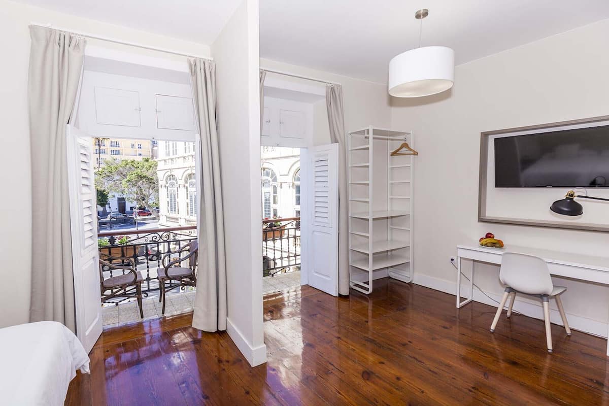 A room with a wooden floor, a bed, a desk with a chair, a TV, and a view of a street and buildings through open doors.