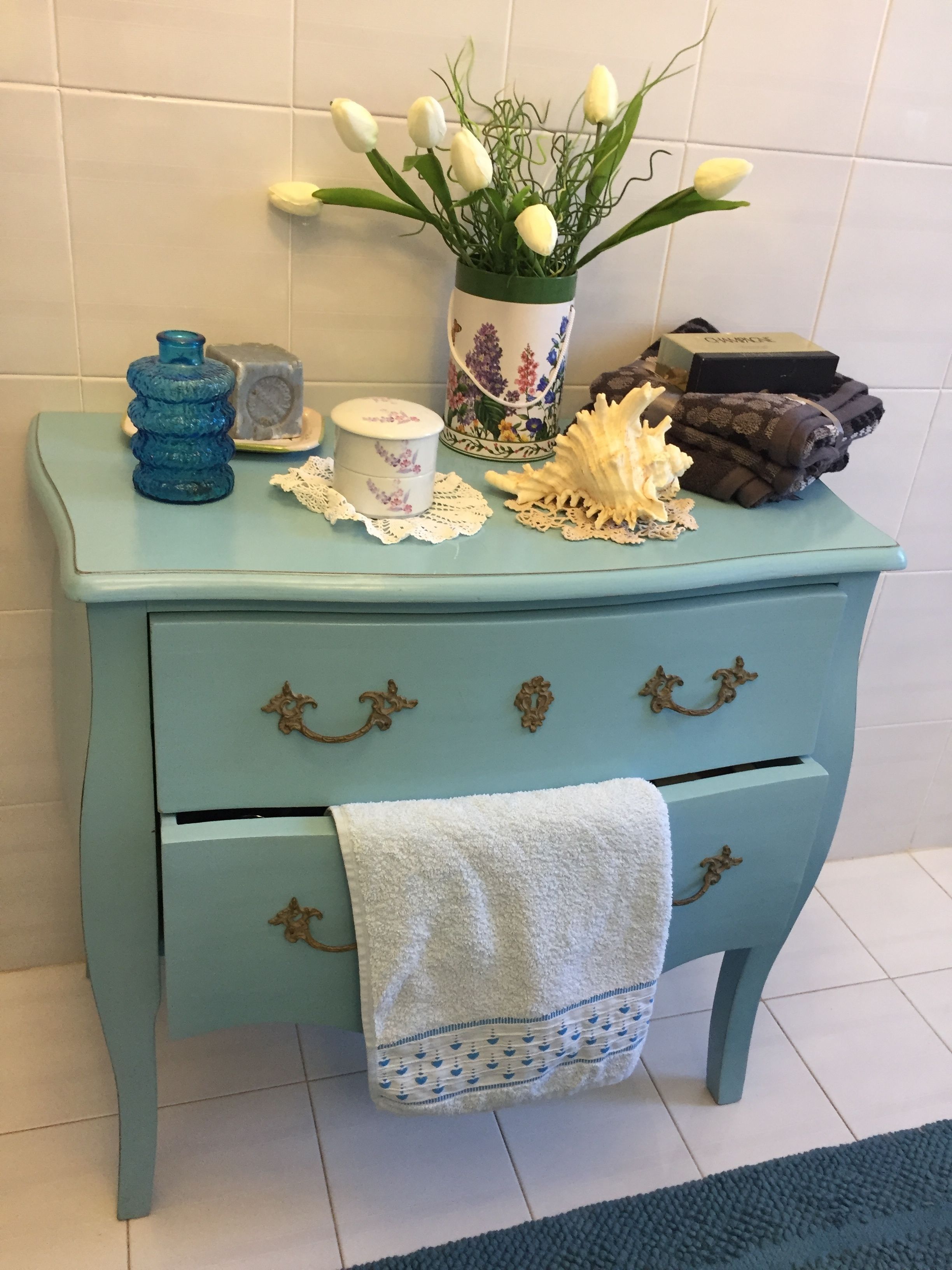 Un aparador azul turquesa con dos cajones, un florero con flores blancas, una botella de vidrio azul, un recipiente blanco pequeño y una concha decorativa.