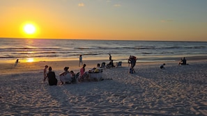 A beach scene with people relaxing and sunbathing, a clear sky, and the sun setting over the ocean.