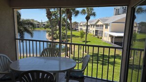 A balcony with a table and chairs, overlooking a body of water and a grassy area with buildings in the background. - 300 OFF APRIL TO NOVEMBER (Naples)