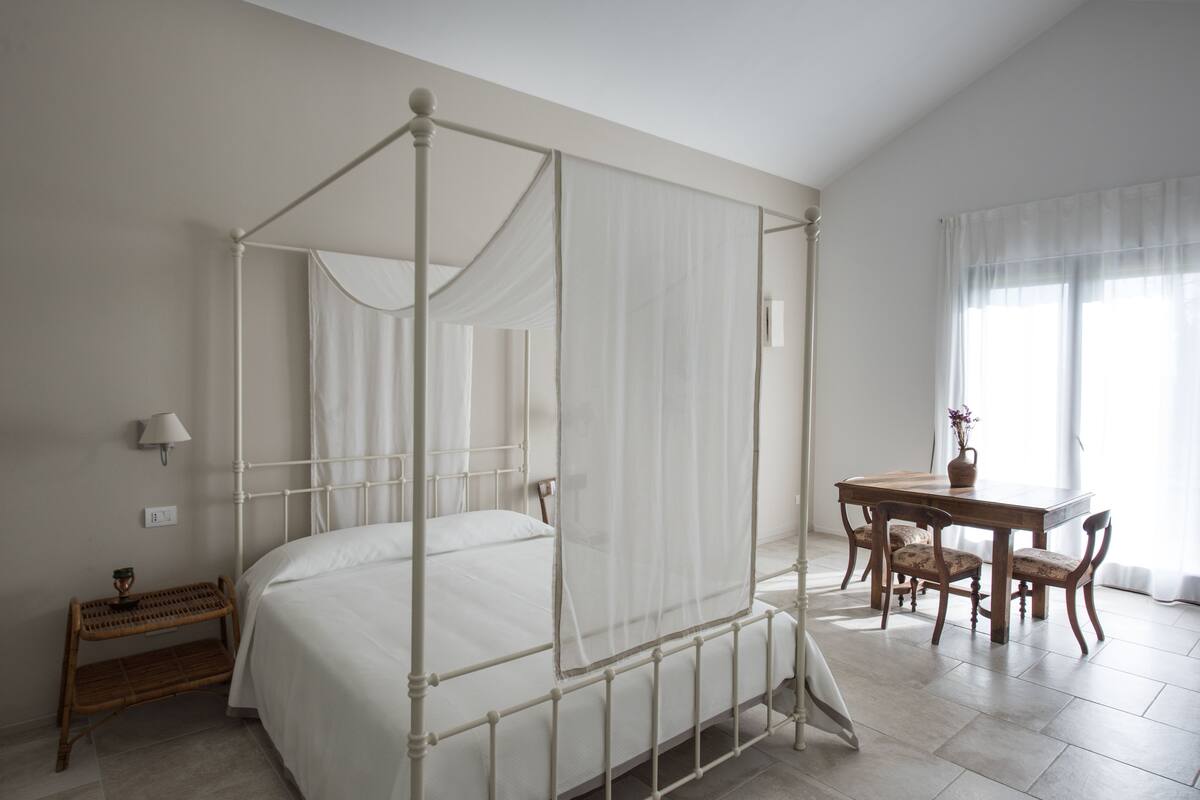 Een slaapkamer met een hemelbed, een houten eettafel met stoelen, een klein nachtkastje met een lamp en een raam met dunne gordijnen.