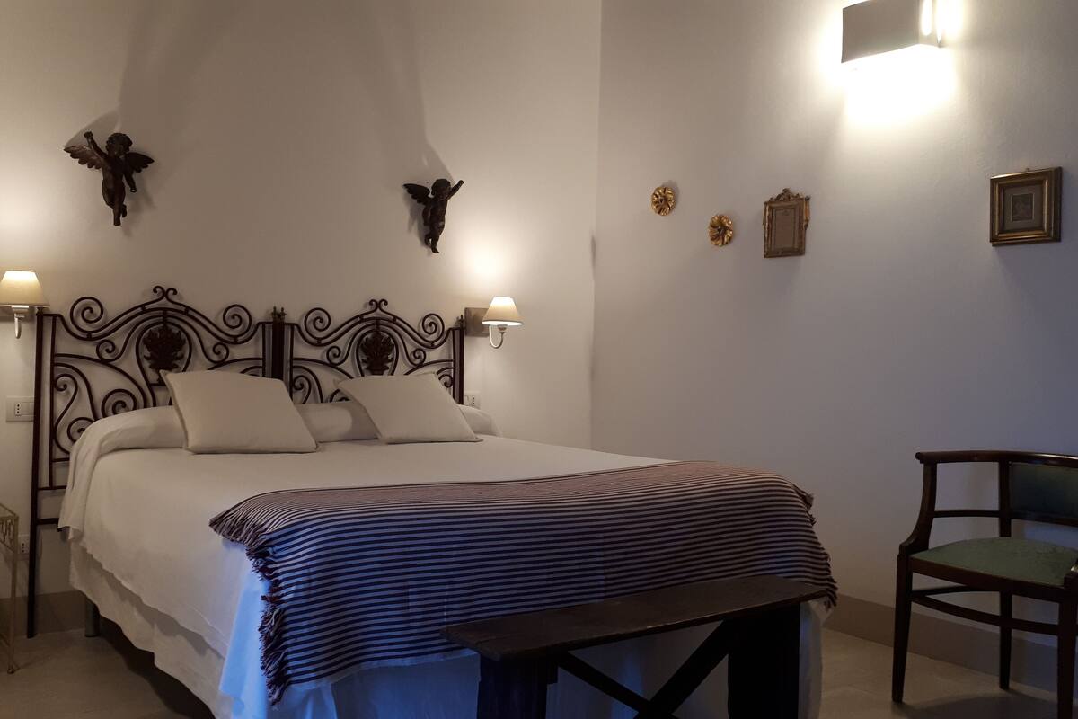 Een net opgemaakt bed met een gestreept dekbed, een houten nachtkastje, een stoel en wandlampen.