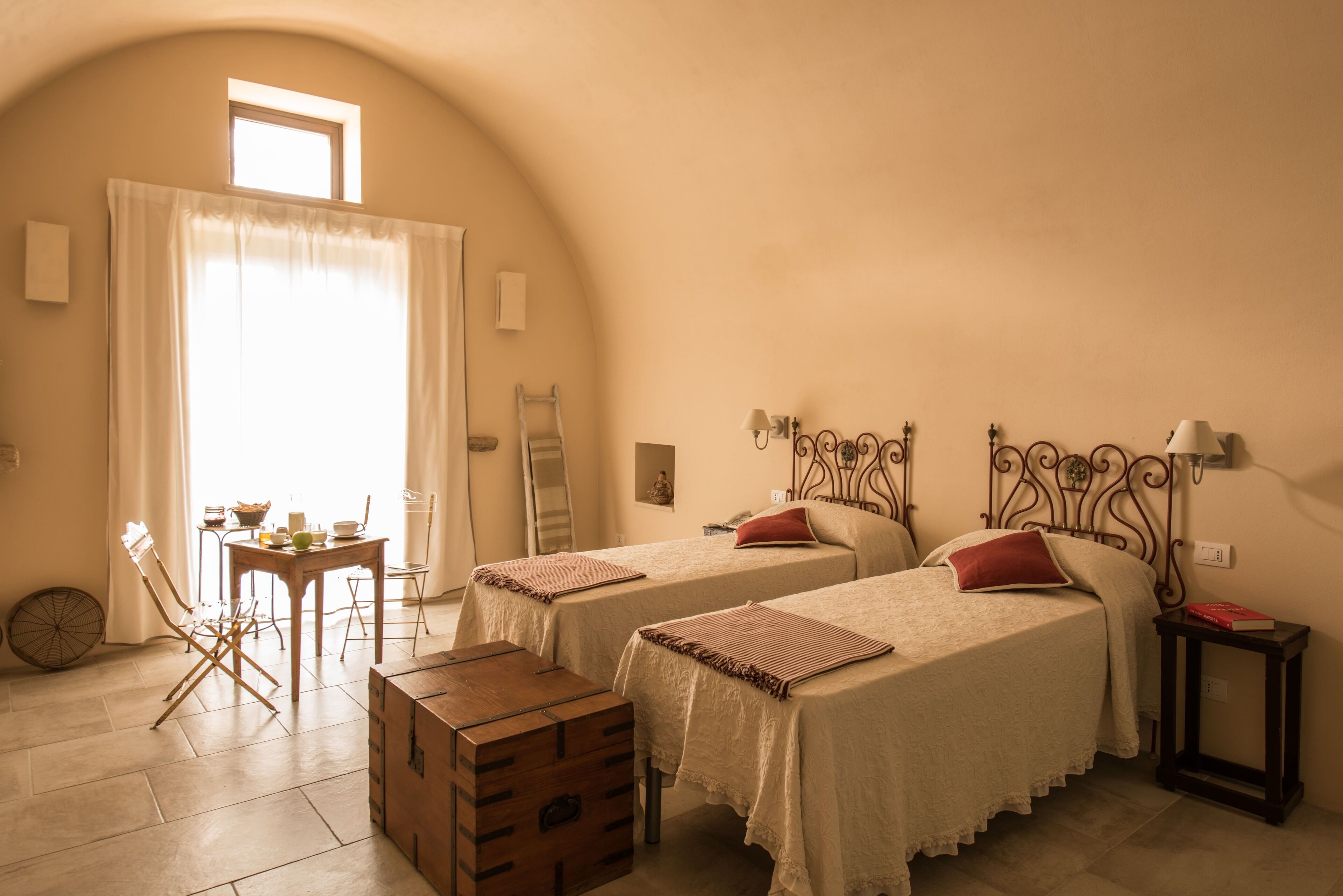 Una habitación con dos camas, un cofre de madera, una mesa con sillas y una ventana con cortinas.