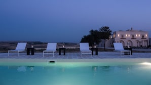 Area piscina con lettini e tavoli, un edificio di grandi dimensioni sullo sfondo e una vista aperta sul paesaggio.