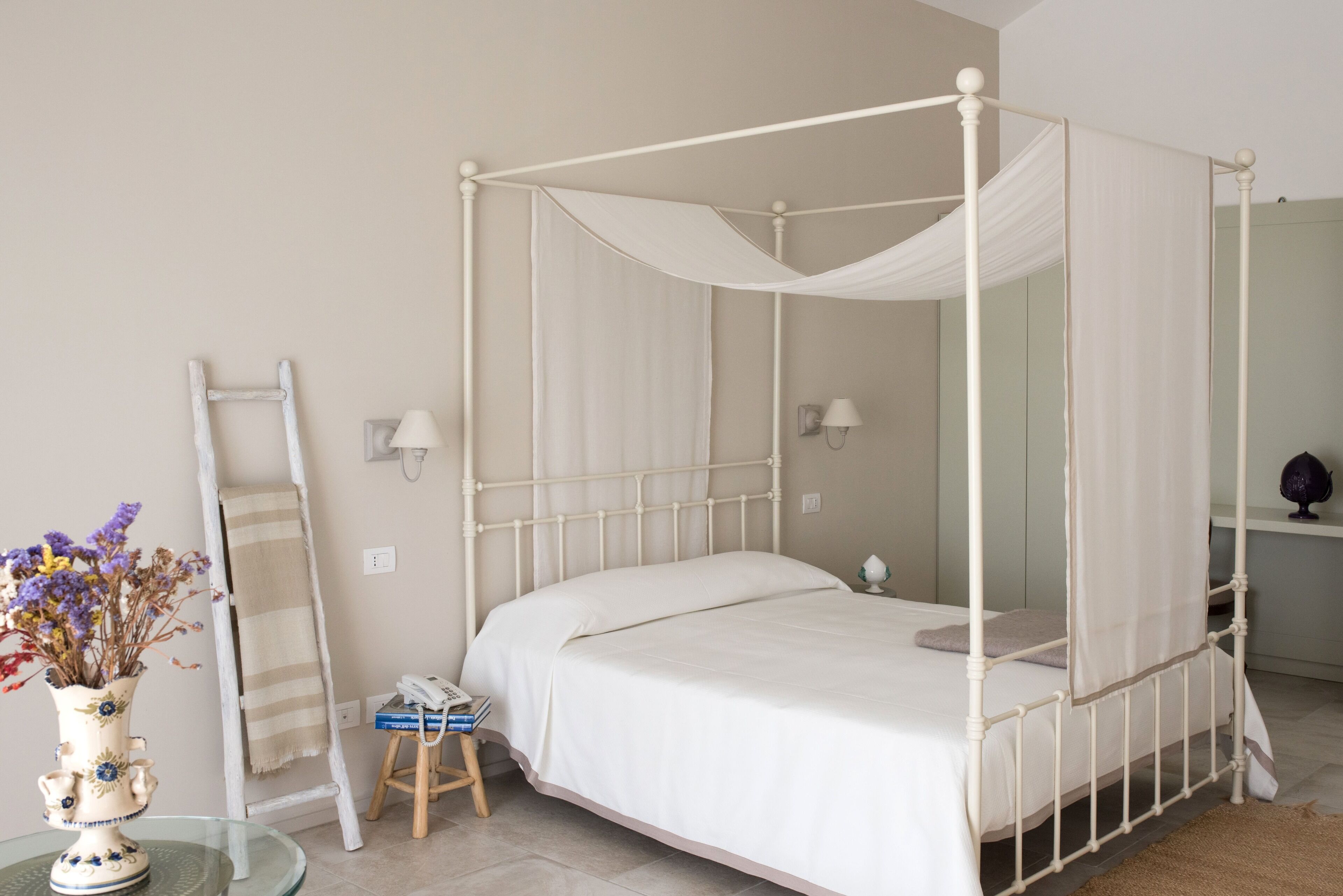 A four-poster bed with a canopy, a bedside table with a lamp, a small stool, and a vase with flowers.