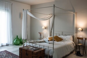 Uma cama de dossel com dossel branco, uma mesa de cabeceira com abajur e um vaso verde no chão.