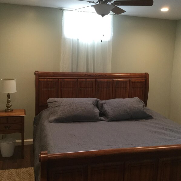A bedroom with a wooden bed, a nightstand with a lamp, a ceiling fan, and a window with curtains. - comfort, luxury, privacy (Amherst)