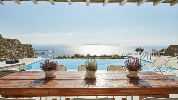 A wooden table with two potted plants, overlooking a pool with a view of the sea and mountains. - Ammos Villas In Houlakia (Mykonos)