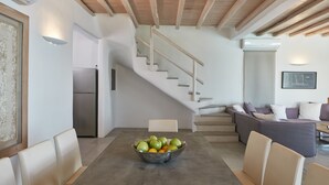 A modern kitchen with a built-in stove, a dining area with a table and chairs, and a staircase leading to a living area with a sofa and cushions. - Ammos Villas In Houlakia (Mykonos)