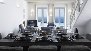 A modern dining area with a long table set for a meal, a television, and a view of the ocean through large windows. - Ammos Villas In Houlakia (Mykonos)