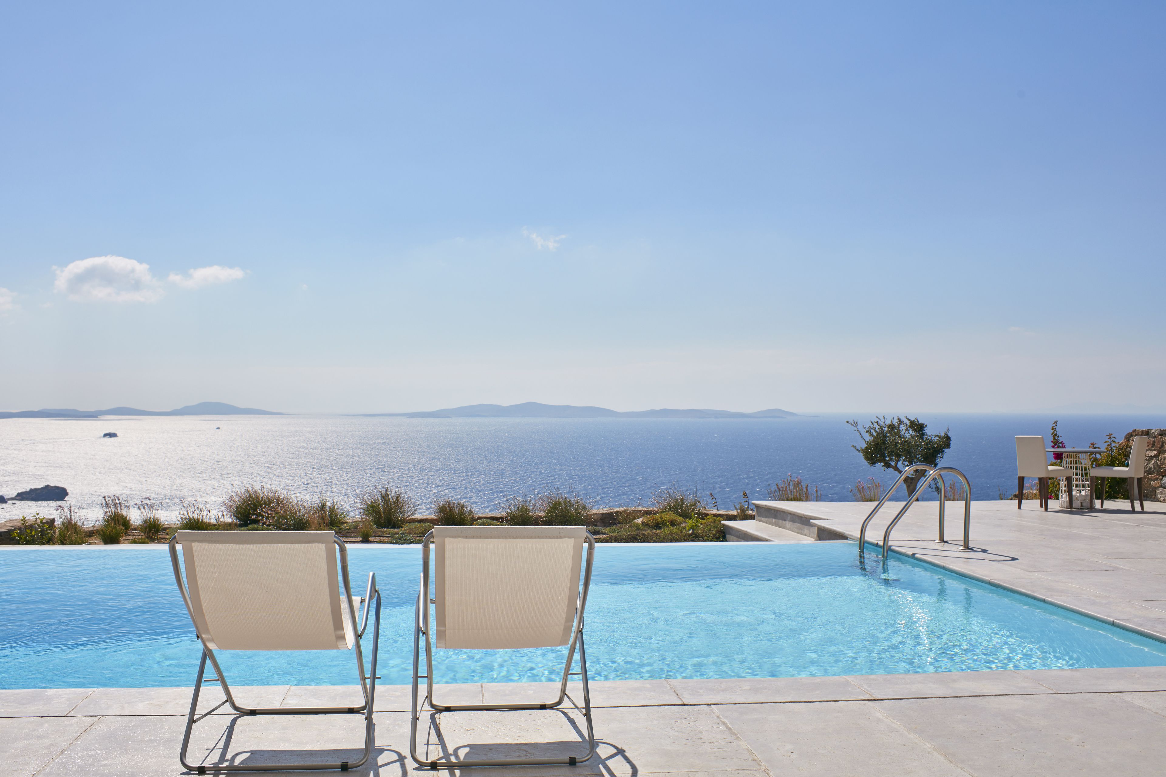 A pool with two chairs facing the sea, a poolside area, and a view of the ocean.