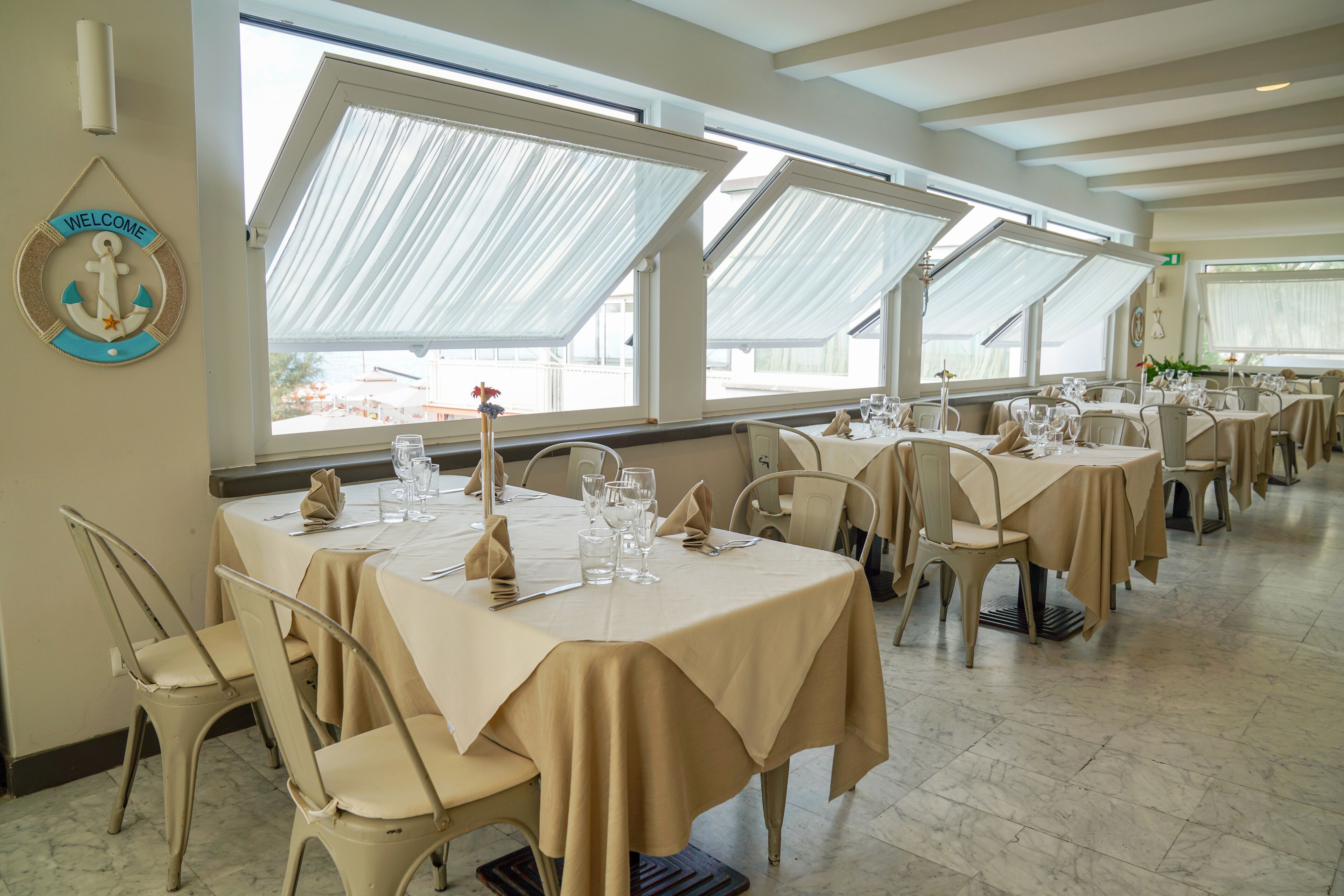 A restaurant with multiple tables set for dining, large windows, and a 'Welcome' sign on the wall.