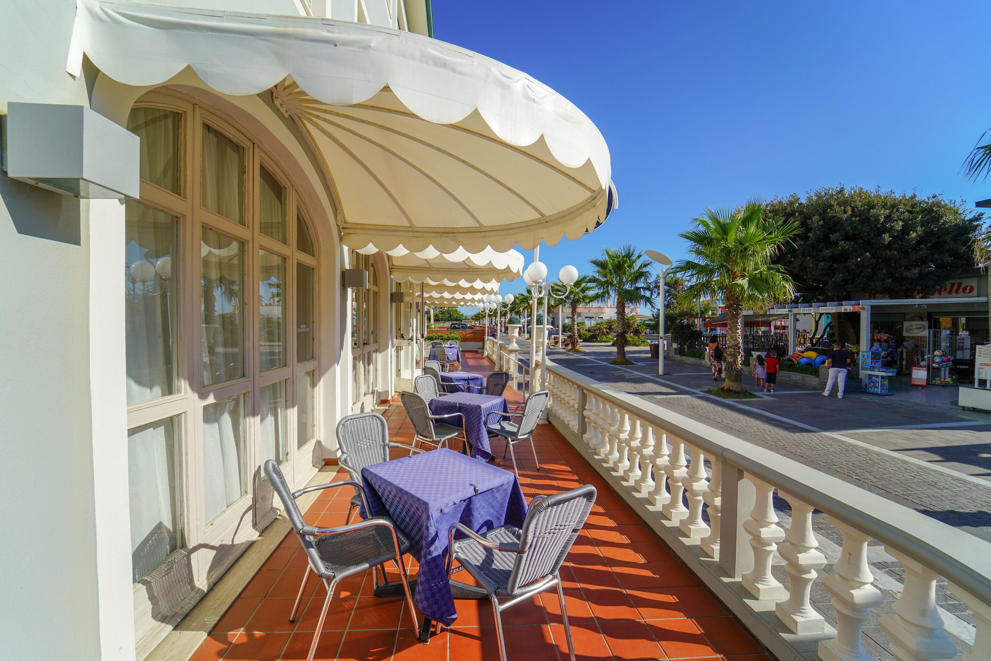 An outdoor cafe with tables and chairs under a large umbrella, a white railing, and palm trees in the background.