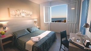 A hotel room with a bed, a desk with a chair, a window with a view of the sea, and a painting on the wall.