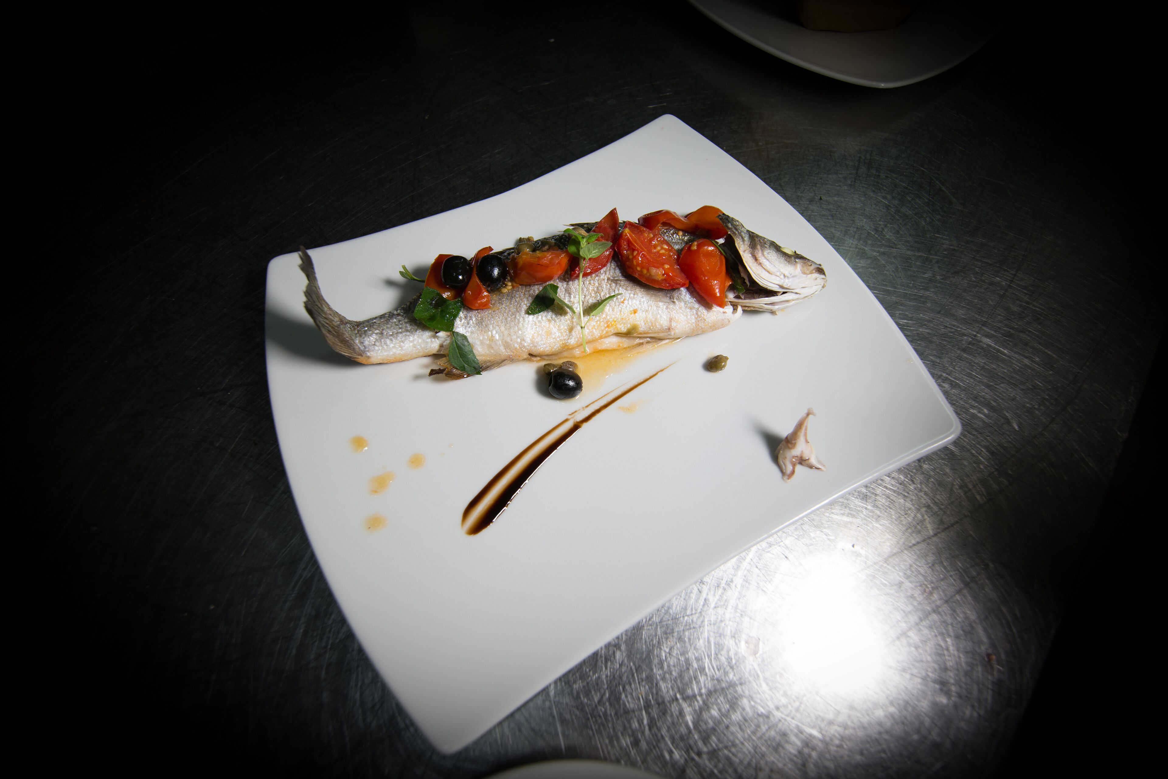Une assiette blanche contenant un plat de poisson, des olives et une sauce, le tout posé sur une surface métallique.