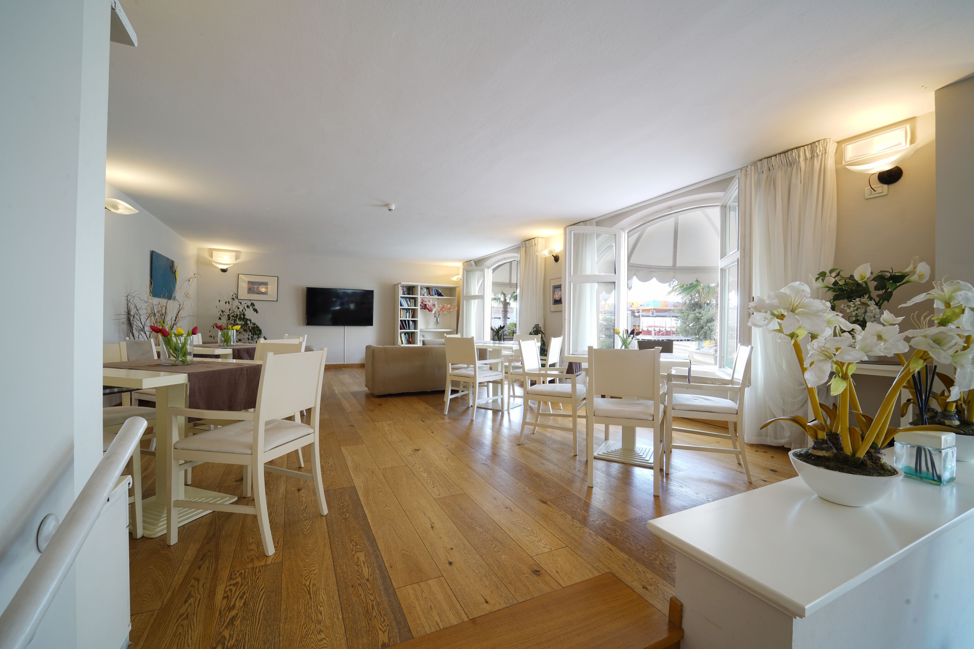 A spacious living room with a wooden floor, white furniture, a dining area, and large windows with curtains.
