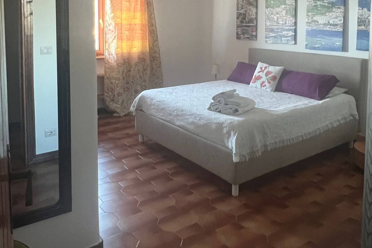 A bedroom with a large bed, wooden flooring, a window with curtains, and wall art.