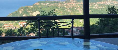 Une terrasse avec une table en verre, un vase vert et un récipient en métal. On aperçoit, au loin, une montagne et une ville côtière.