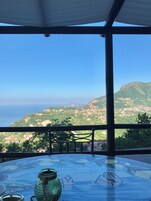 Una terraza con una mesa de vidrio, un jarrón verde y un recipiente de metal. Se divisa una montaña y un pueblo costero al fondo.