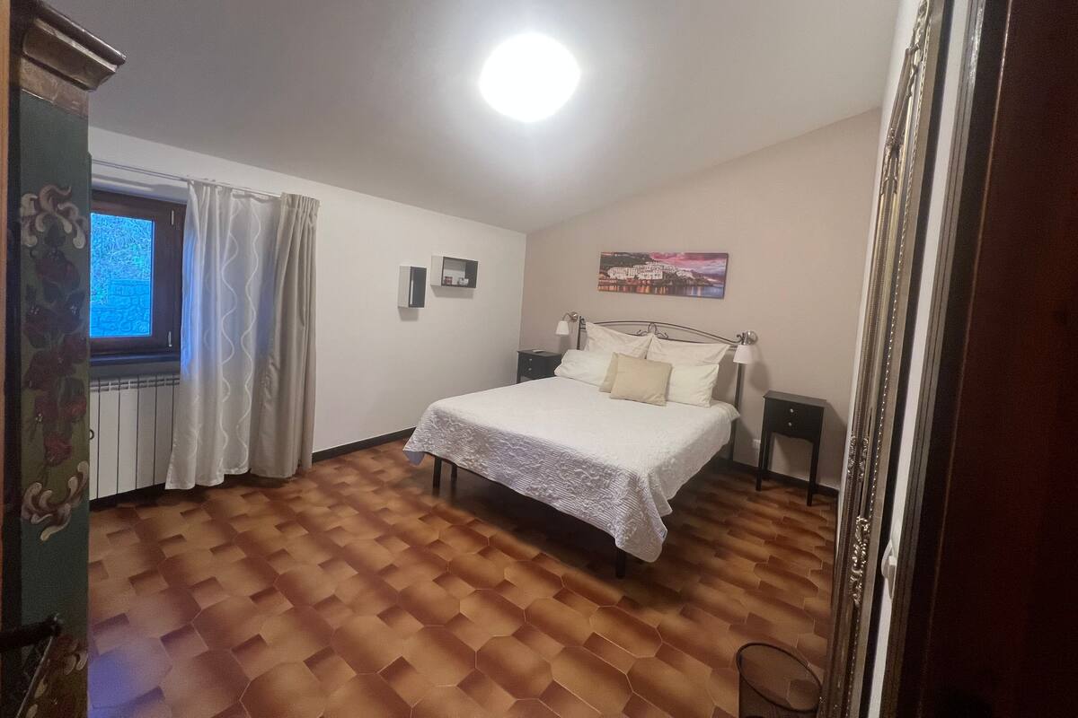 A bedroom with a bed, a nightstand, a window with curtains, a radiator, and a painting on the wall.