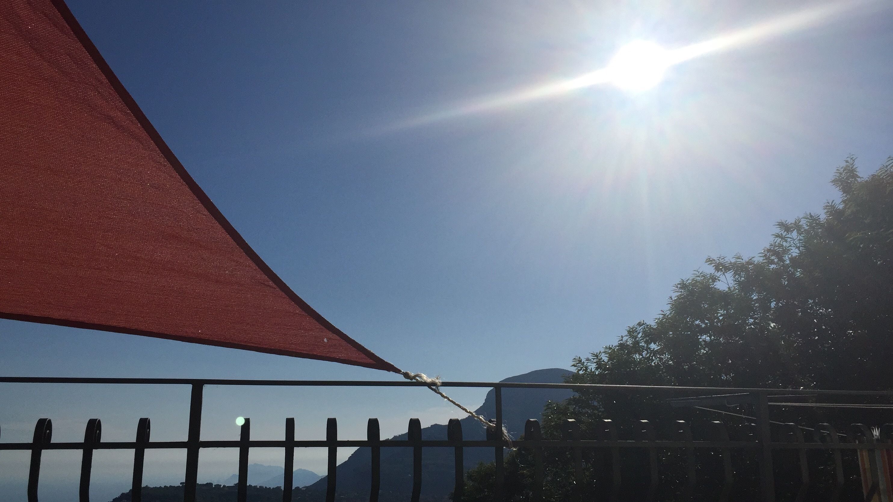Cielo despejado con el sol brillando intensamente, una sombra triangular roja y una valla de metal.
