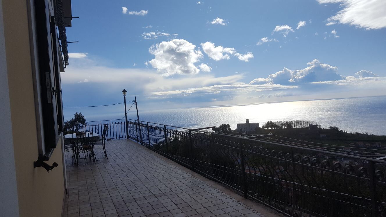 A balcony with a tiled floor, a table with chairs, and a view of the sea and buildings in the distance.