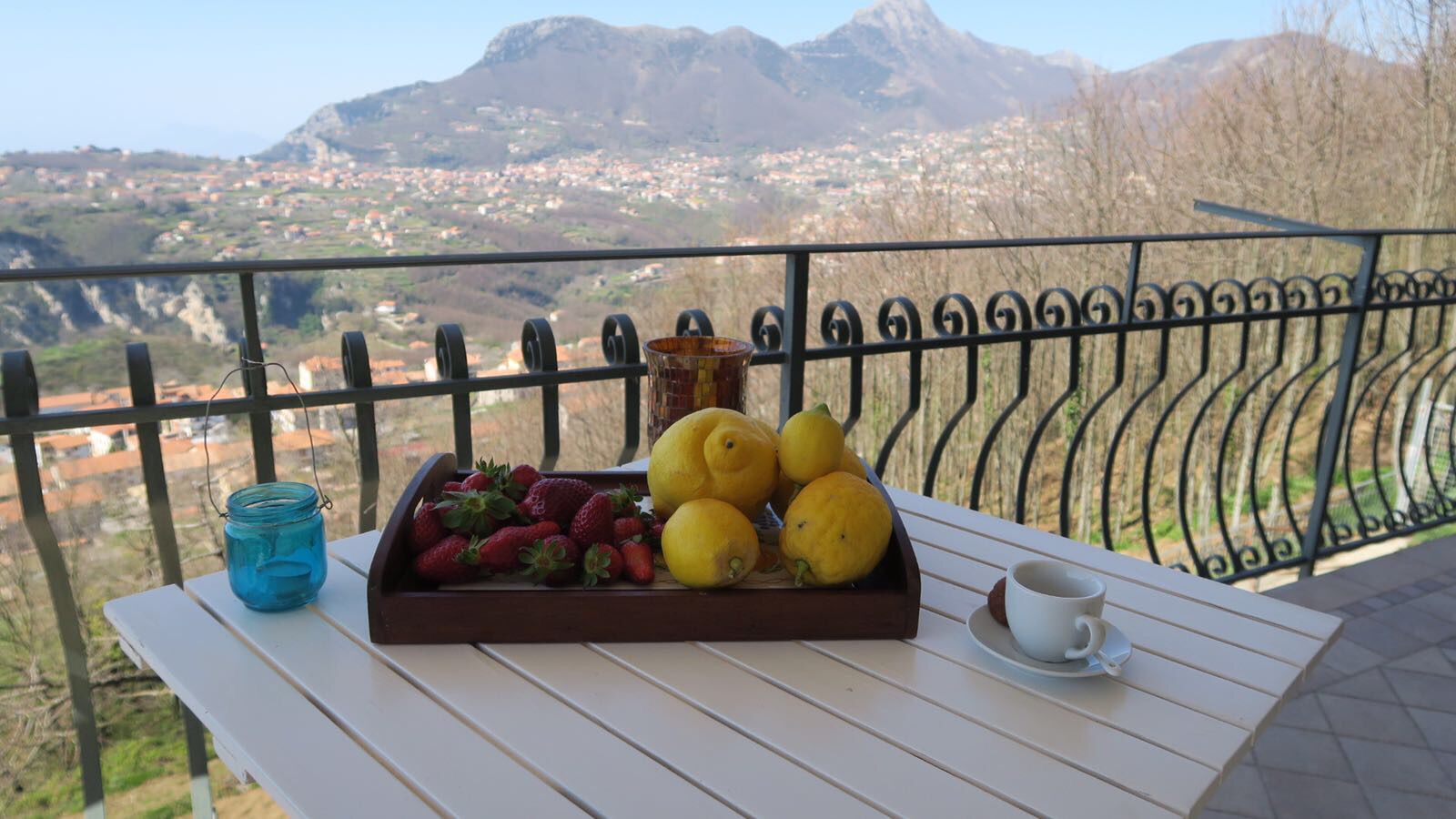 A table with a tray of strawberries and lemons, a blue jar, and a cup on a balcony with a mountain view.
