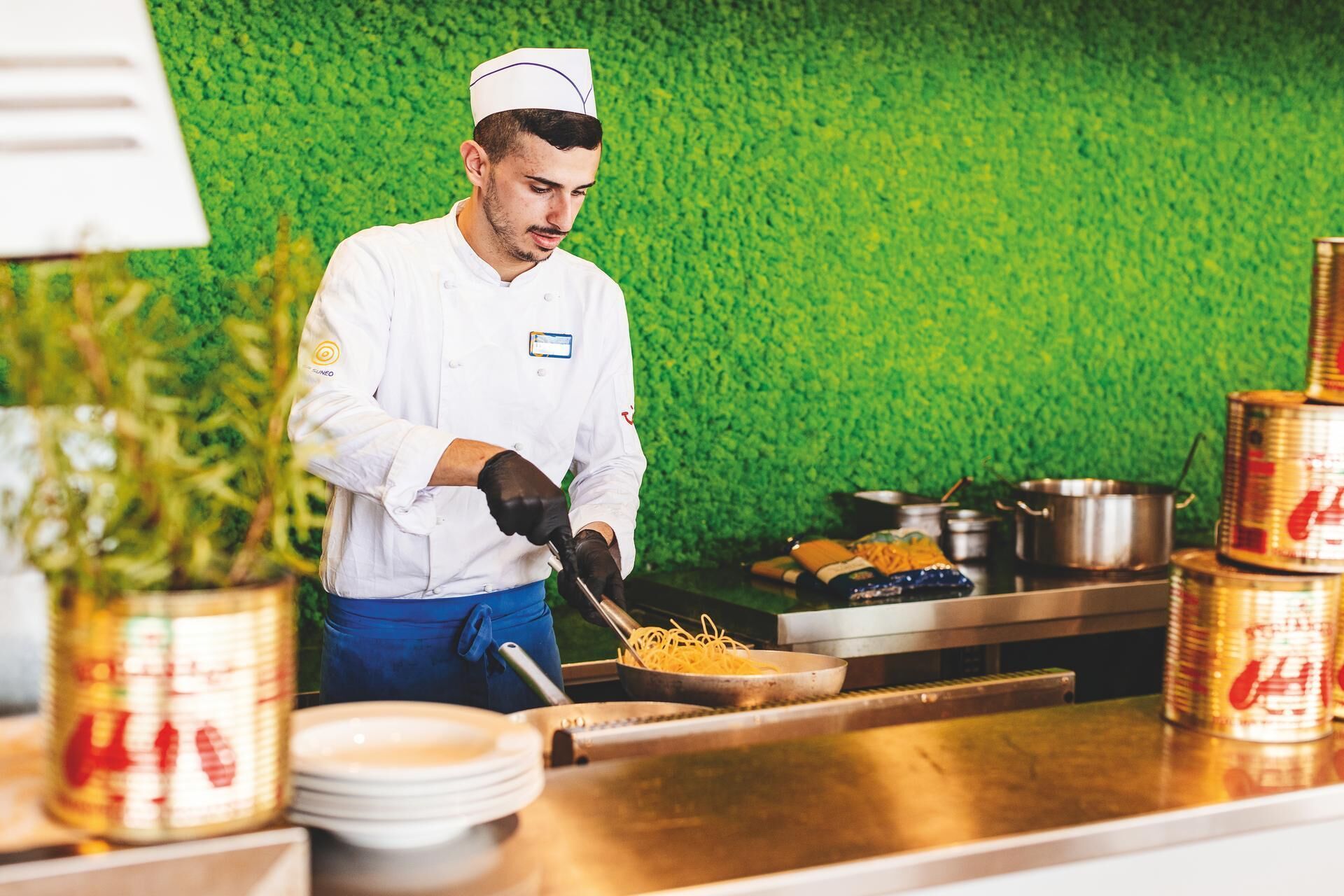 A chef in a white uniform preparing food in a kitchen with a green wall and stainless steel appliances.