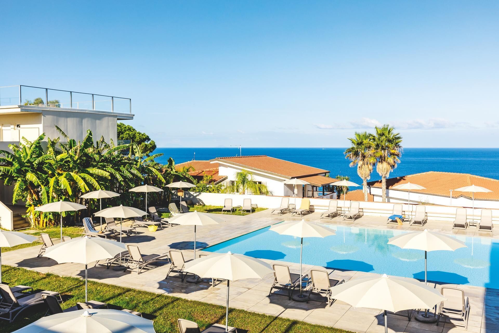 Uma área de piscina com espreguiçadeiras e guarda-sóis, vista clara para o oceano e um edifício com telhado em azulejos.