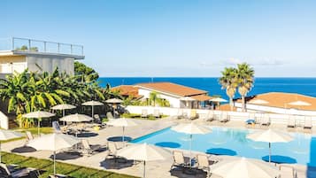 Uma área de piscina com espreguiçadeiras e guarda-sóis, vista clara para o oceano e um edifício com telhado em azulejos.