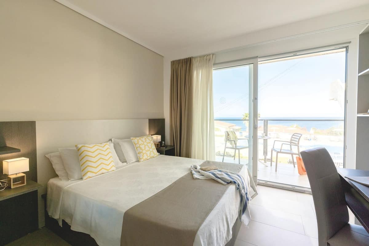 A hotel room with a bed, a desk, a chair, a lamp, and a view of the sea through a sliding door.