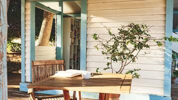 Una mesa de madera con un libro y una taza, un banco y un árbol frente a un edificio blanco con detalles azules.