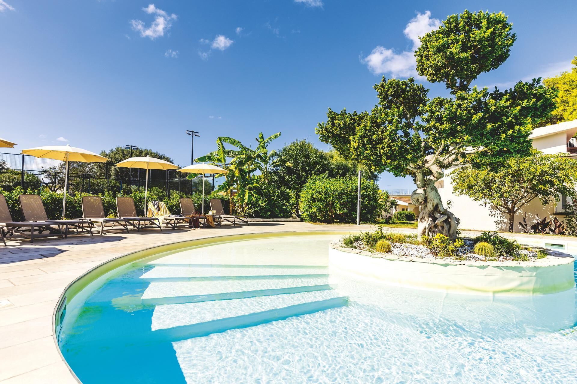 A pool area with lounge chairs and umbrellas, surrounded by greenery and a statue.
