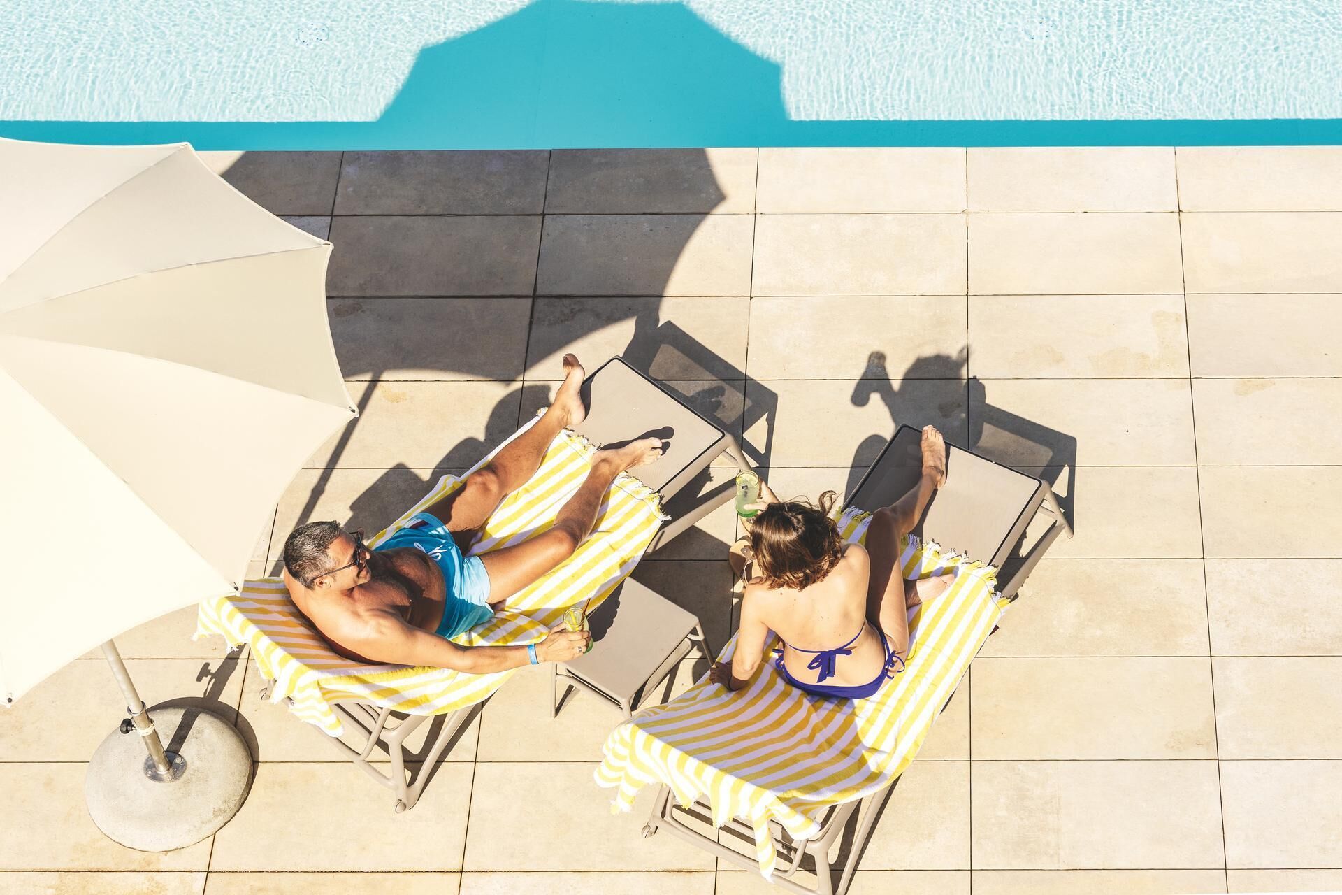 Two people sunbathing on lounge chairs by a pool with a tiled floor and a large umbrella.