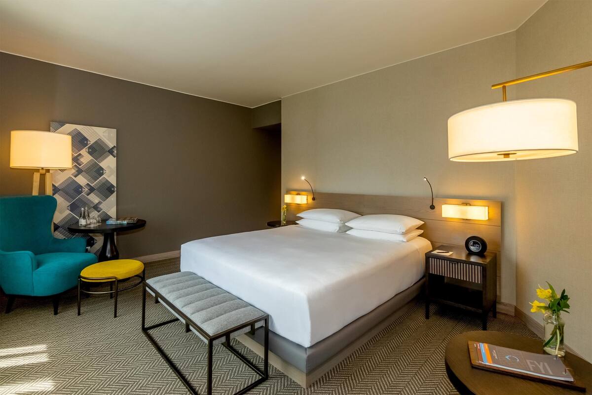 Una habitación de hotel con una cama grande, un escritorio, una silla, una mesita pequeña y una mesita de noche con lámpara.