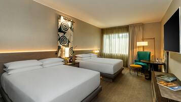 A hotel room with two beds, a desk with a chair, a lamp, and a wall-mounted artwork.