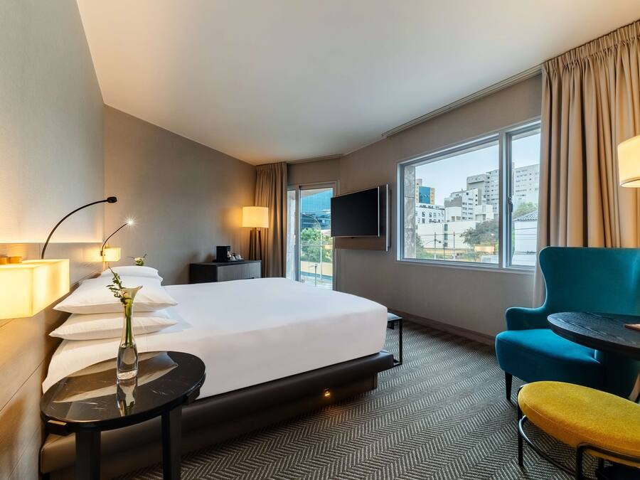 Habitación de hotel con cama, mesita de noche, lámpara, sillón y vistas a la ciudad a través de amplios ventanales.