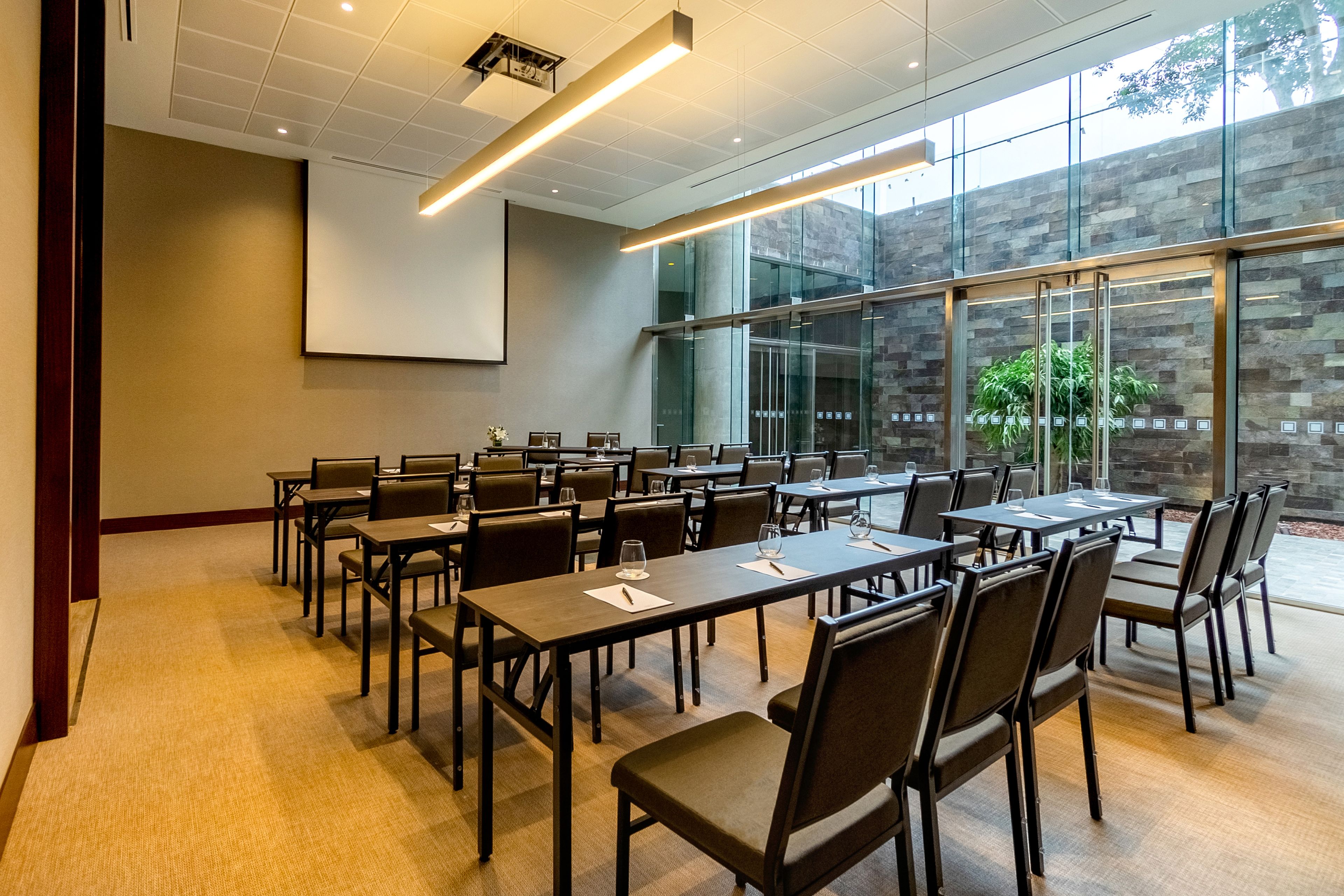 Un salón de conferencias con varias mesas y sillas, ventanas amplias y una pantalla para proyector.