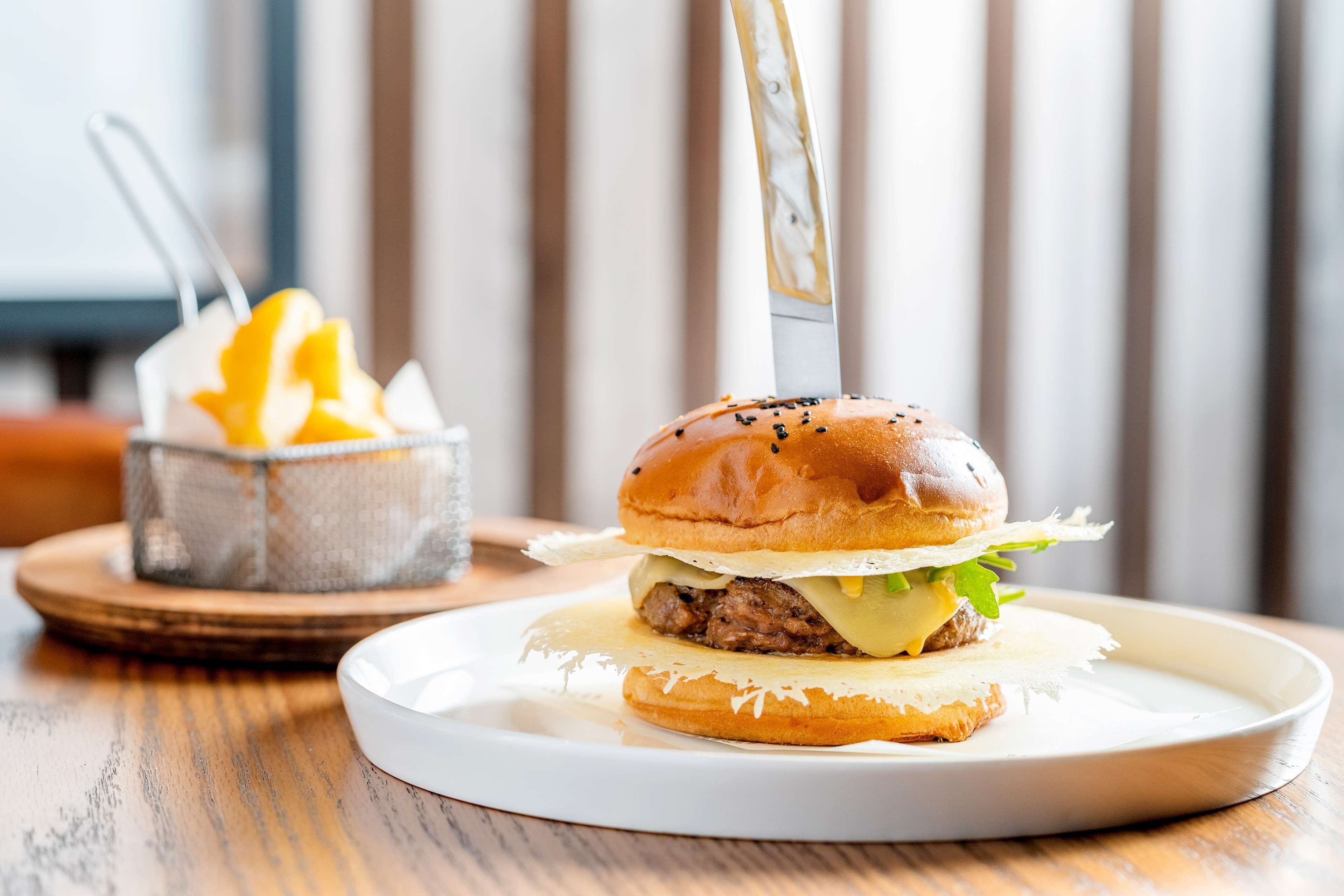 A burger on a white plate with a knife on top, accompanied by a small bowl of fries and a drink in the background.