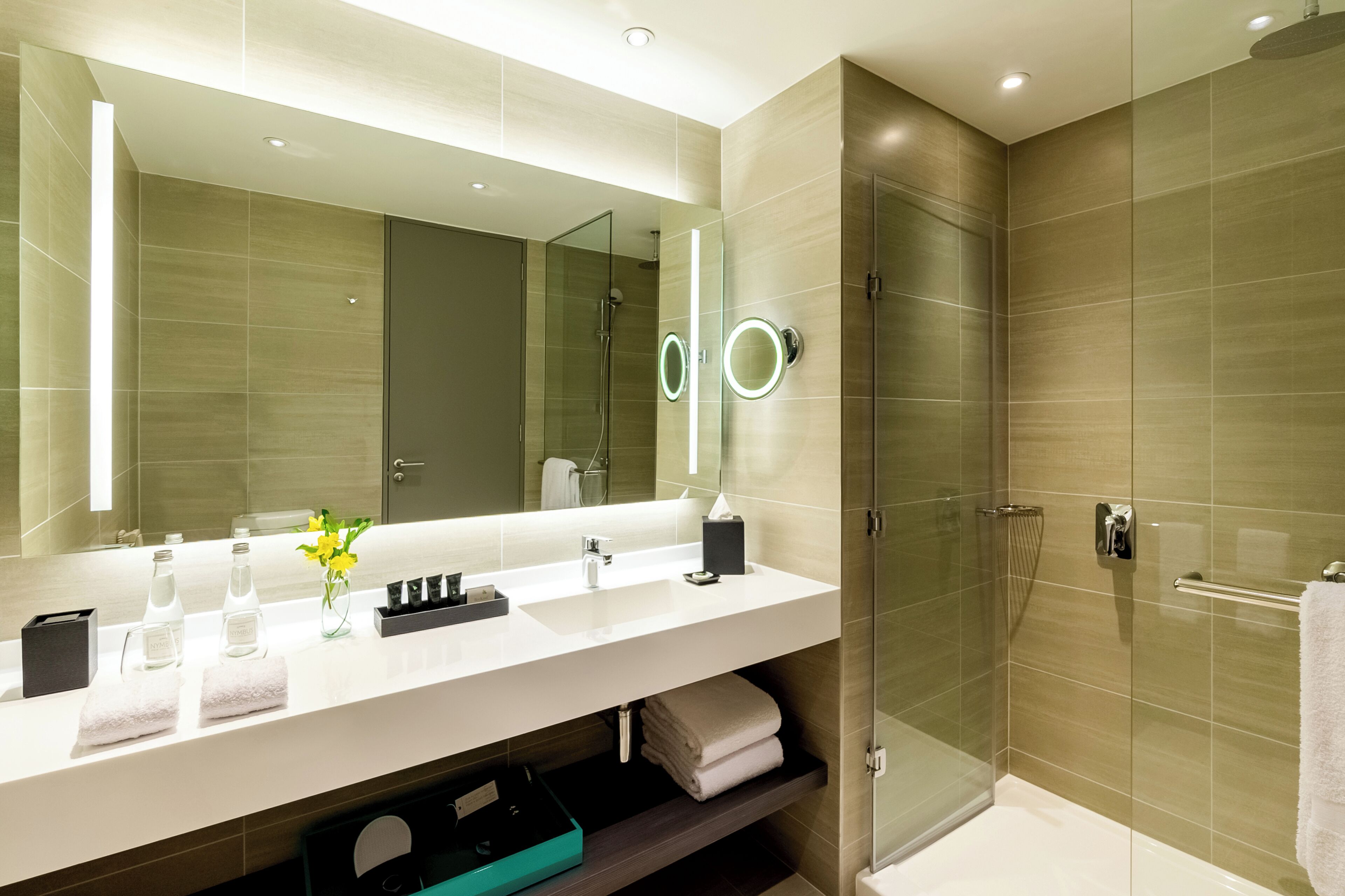 Un baño moderno con un espejo grande, cabina de ducha de vidrio y tocador blanco con artículos de aseo.