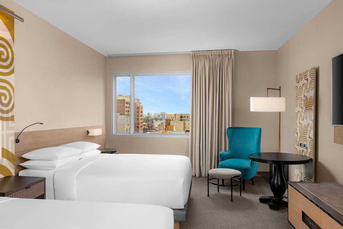 Habitación de hotel con dos camas, un sillón verde azulado, una mesita, un televisor y una ventana con vista a la ciudad.