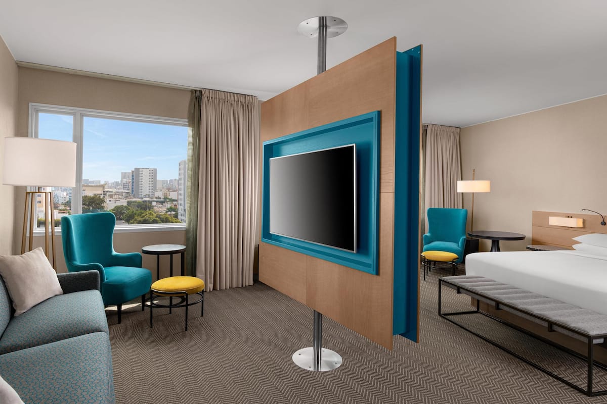 Habitación de hotel con una televisión grande, una cama, un sofá, una silla, una mesita y vistas a la ciudad.