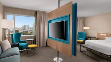 A hotel room with a large TV, a bed, a sofa, a chair, a small table, and a view of the city.