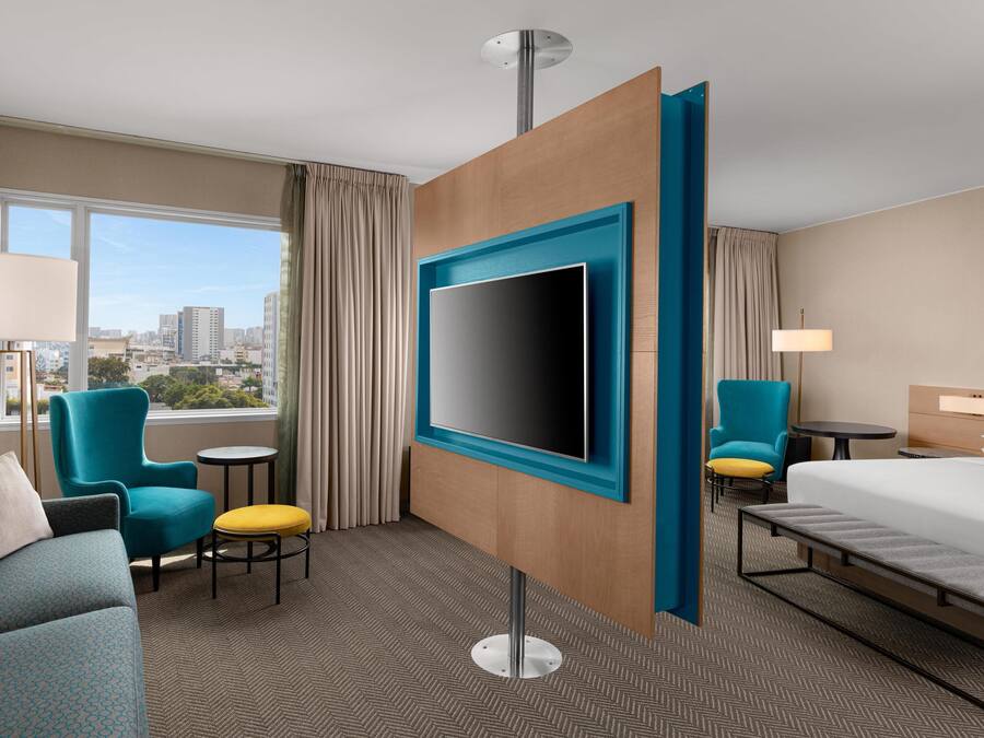 A hotel room with a large TV, a bed, a sofa, a chair, a small table, and a view of the city.