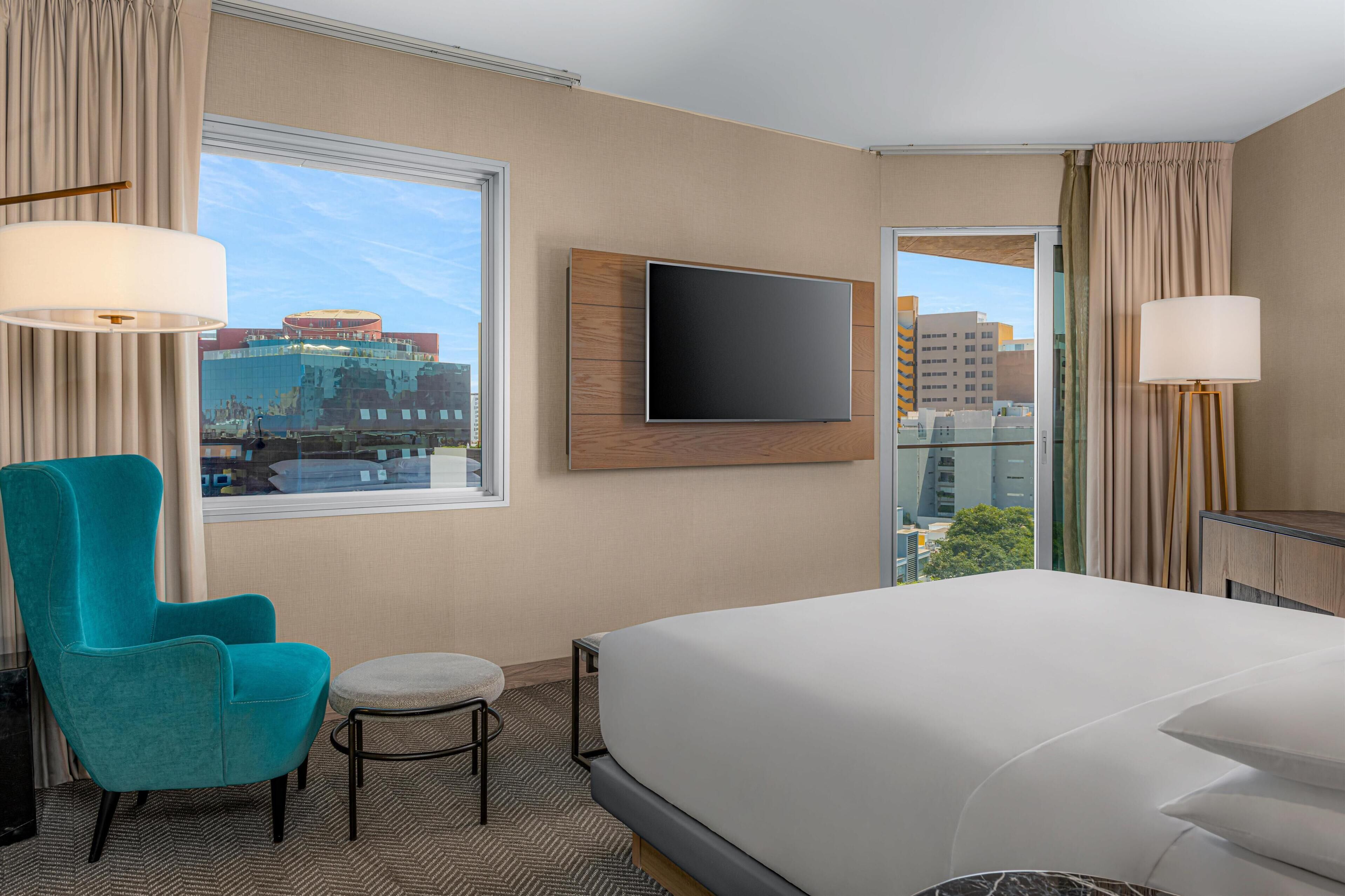 Una habitación de hotel con una cama grande, un televisor en la pared, un sillón azul verdoso y un taburete pequeño.